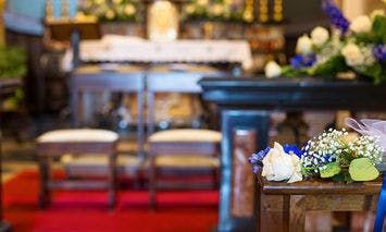 A floral bouquet rests on a wooden pew in a decorated church interior, featuring an altar adorned with flowers and framed by two prayer benches on a red carpet.