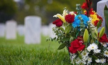 A bouquet of vibrant flowers is placed in front of a white gravestone, surrounded by grassy terrain and additional gravestones in the background of a cemetery. No text is present.