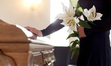 A person in black holds white lilies while touching a wooden casket, situated in a softly-lit, somber room.
