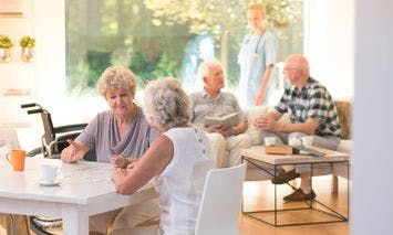 Elderly individuals are seen sitting and conversing around a well-lit, cozy room with large windows. A caregiver stands in the background, while two women work on a puzzle at a table.