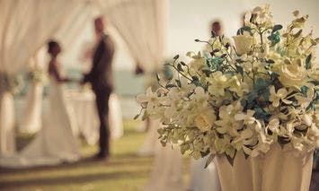 A bouquet of white and blue flowers sits in the foreground, with a blurred wedding ceremony taking place in a grassy outdoor setting in the background.